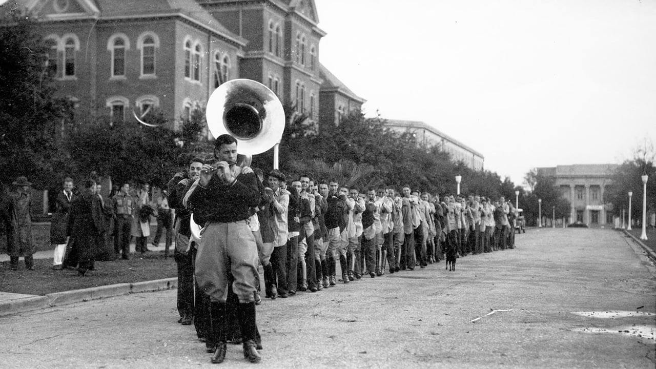 Black-and-white photo of Texas A&M students marching in a line, led by a sousaphone player.