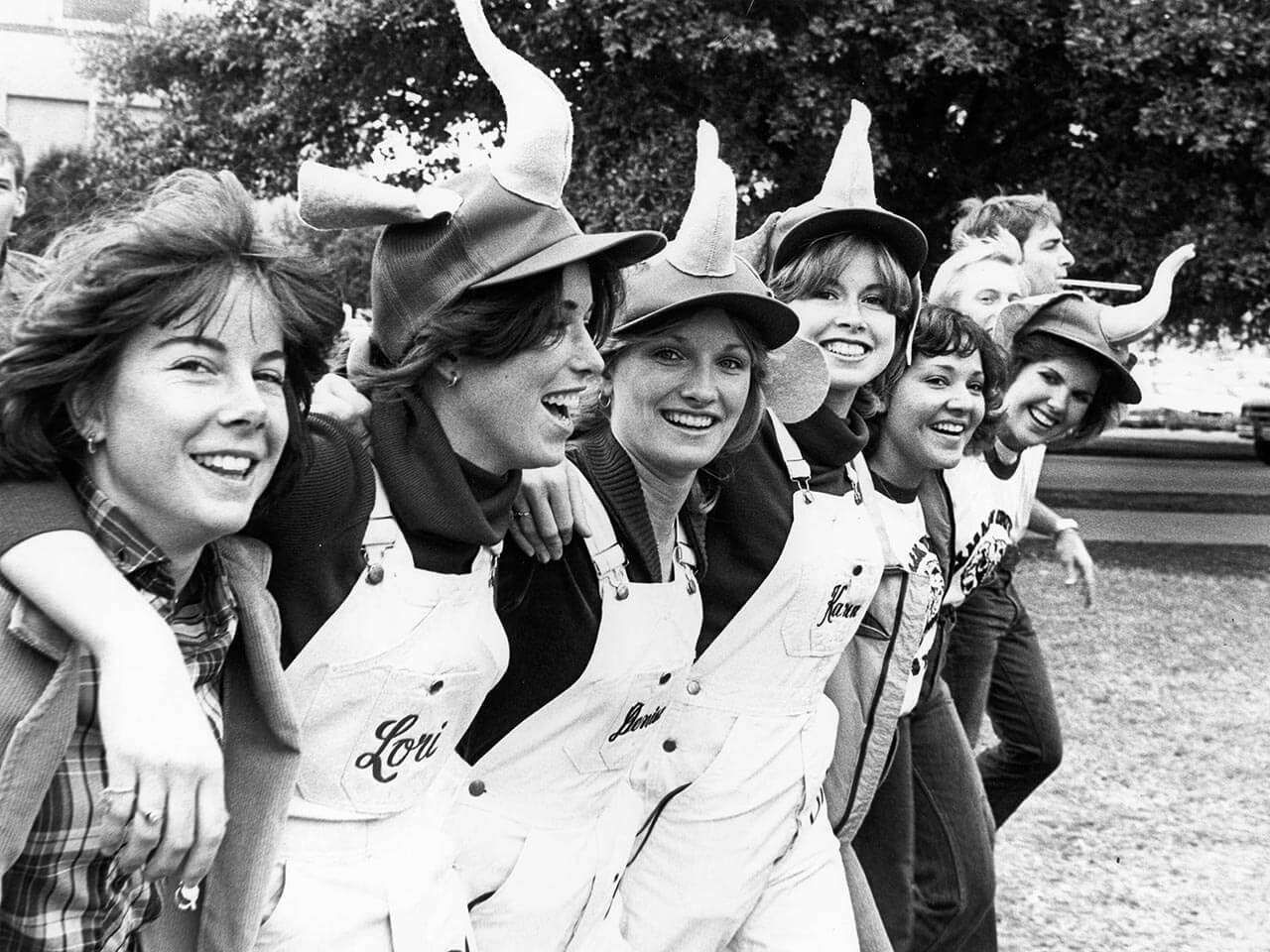 Black-and-white photo of Texas A&M students in elephant hats and T-shirts with arms linked during Elephant Walk.